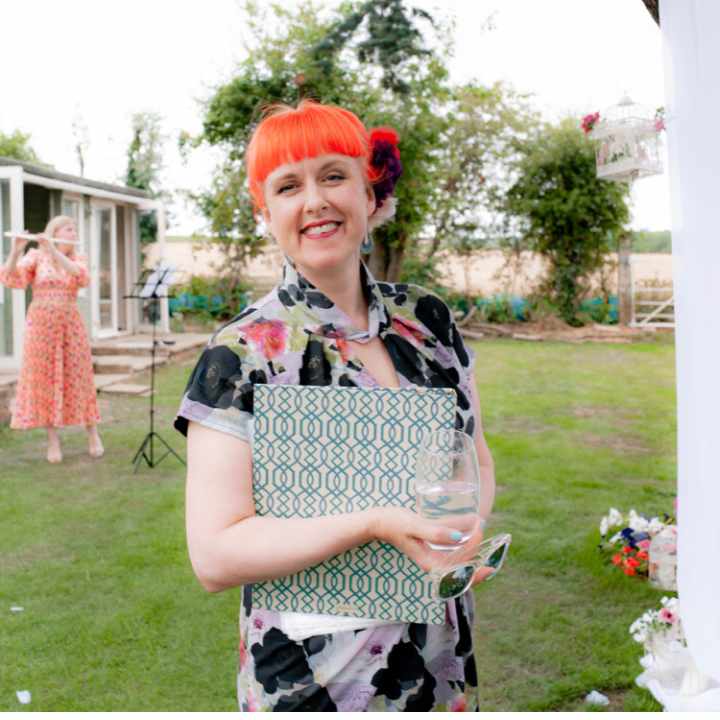 Civil Funeral Celebrant Jess May smiling at the camera holding a patterned folder. She is outside and has a dress with short sleeves and orange hair.