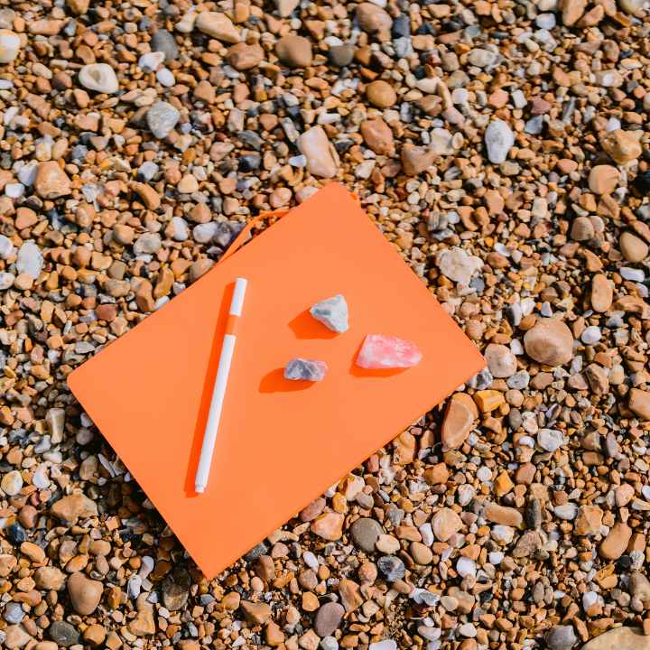 an orange notebook with 3 crystals and a pen on the beach
