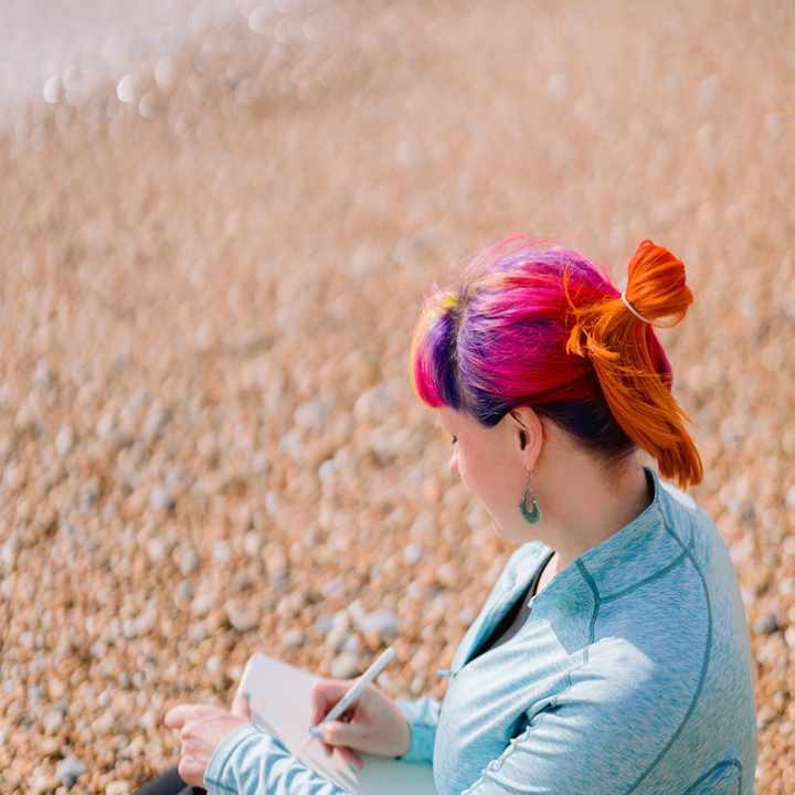 Jess May sitting on the beach writing. She is wearing a turquoise top and has her hair up . her hair is blue, pink and yellow and orange