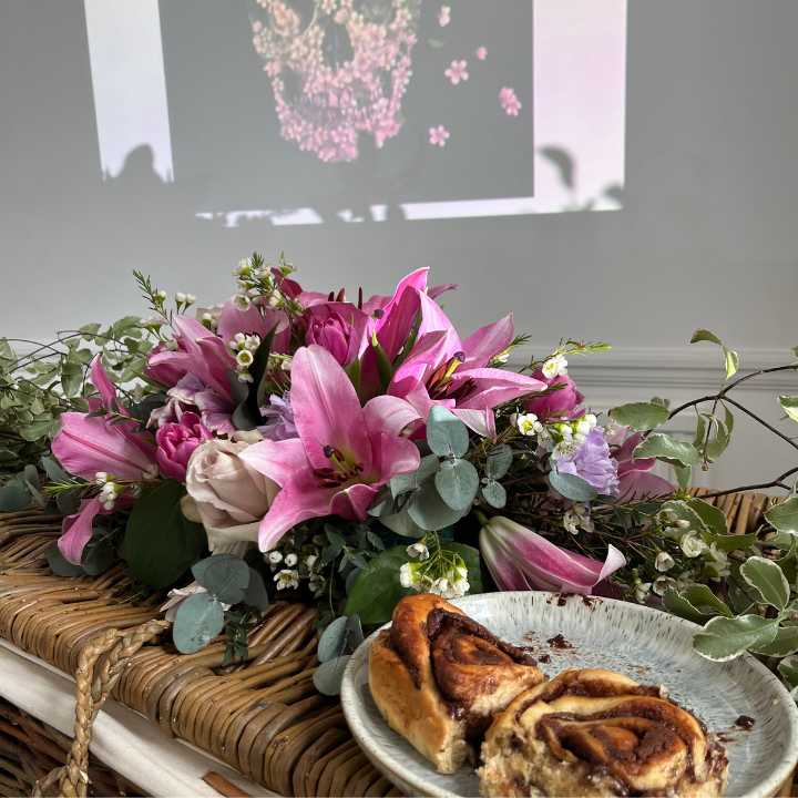 A pink floral funeral tribute made from lilies with a cinnamon bun on a plate.