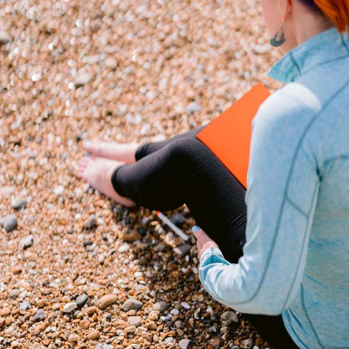 Jess May Brighton Funeral Celebrant on a beach wearing turquoise top and black leggings and holding an orange notebook for creating DIY funerals.
