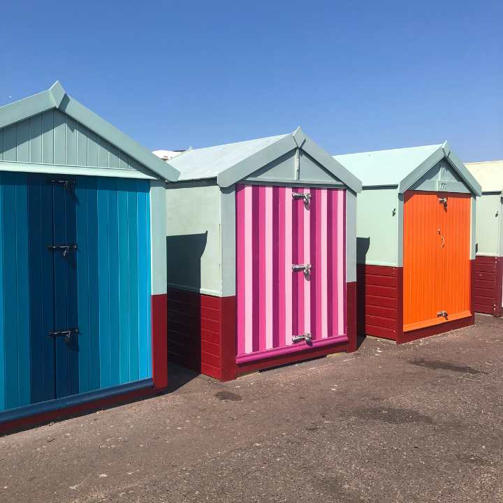 Orange, pink and turquoise beach huts. The blue/turquoise and the pink have vertical stripes. The wooden rooves are pale turquoise. they have a sea side feel like a holiday.