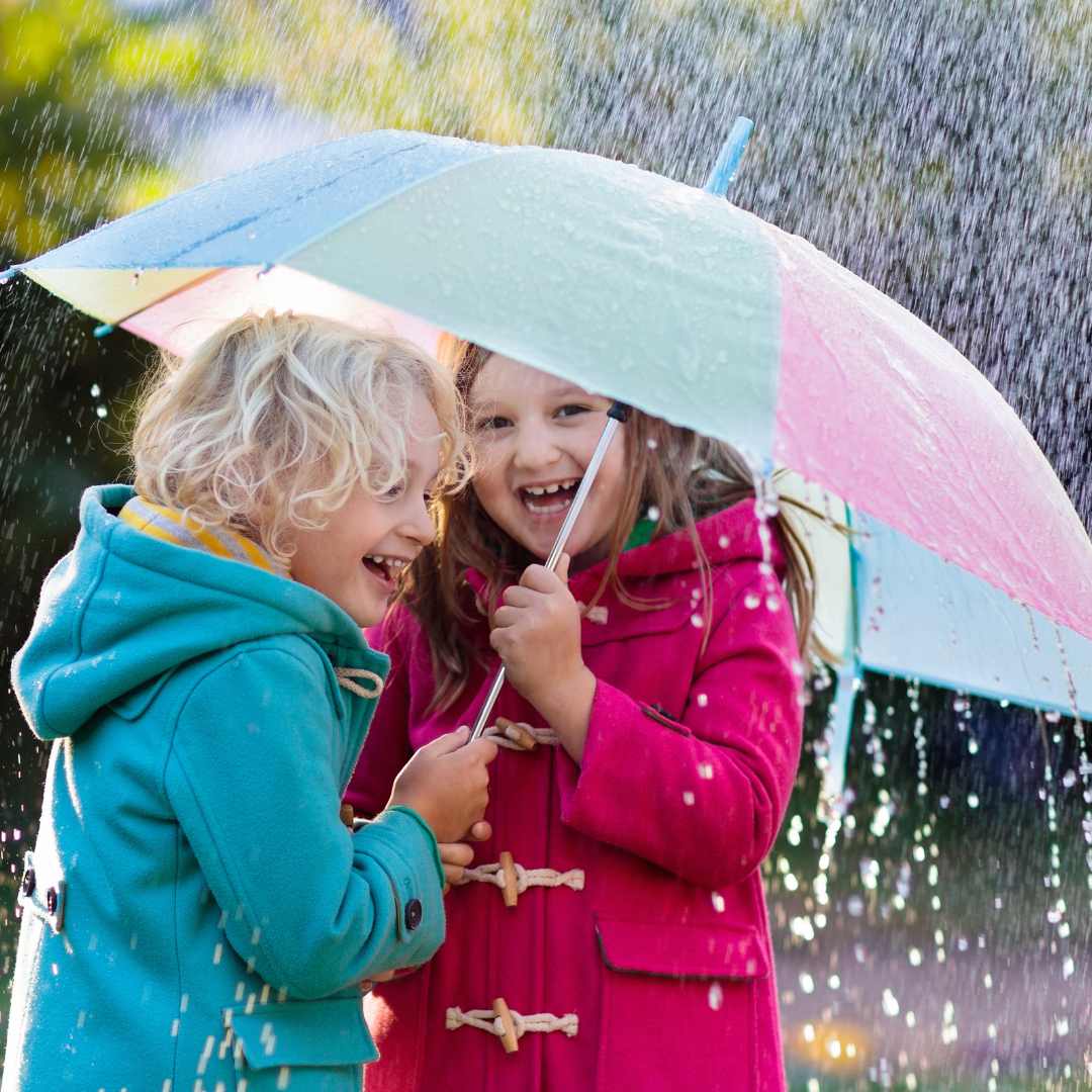 Two children under a pastel coloured rainbow umbrella, the boy has white hair and a turquoise coat and the girl is wearing pink. They are laughin.