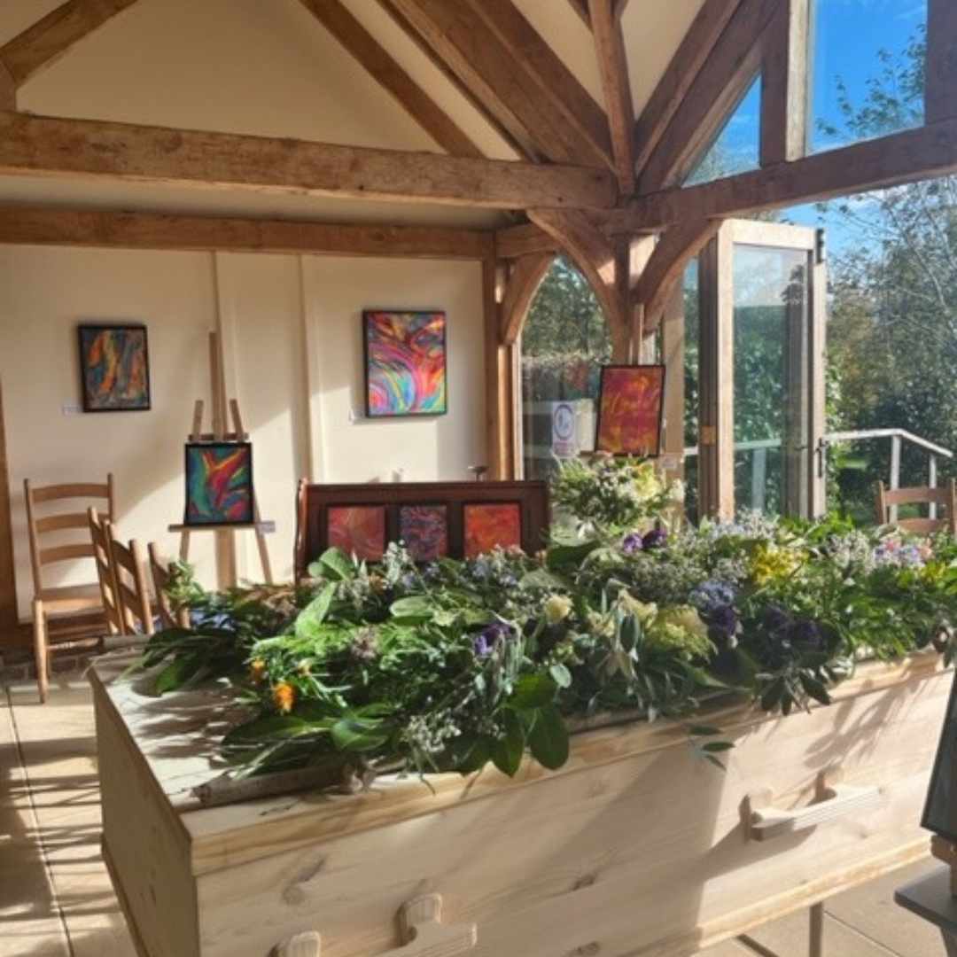 Funeral Venue Clayton Woods Natural Burial Ground. Light filled with timber frame building, paintings in the background and a coffin with flowers on top