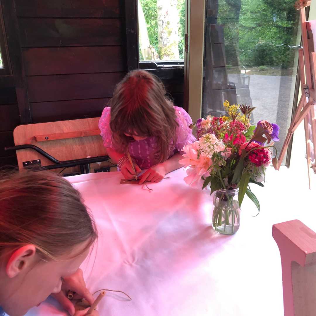 At a Living Wake two children are drawing with a bunch of flowers on the table. The light i pink