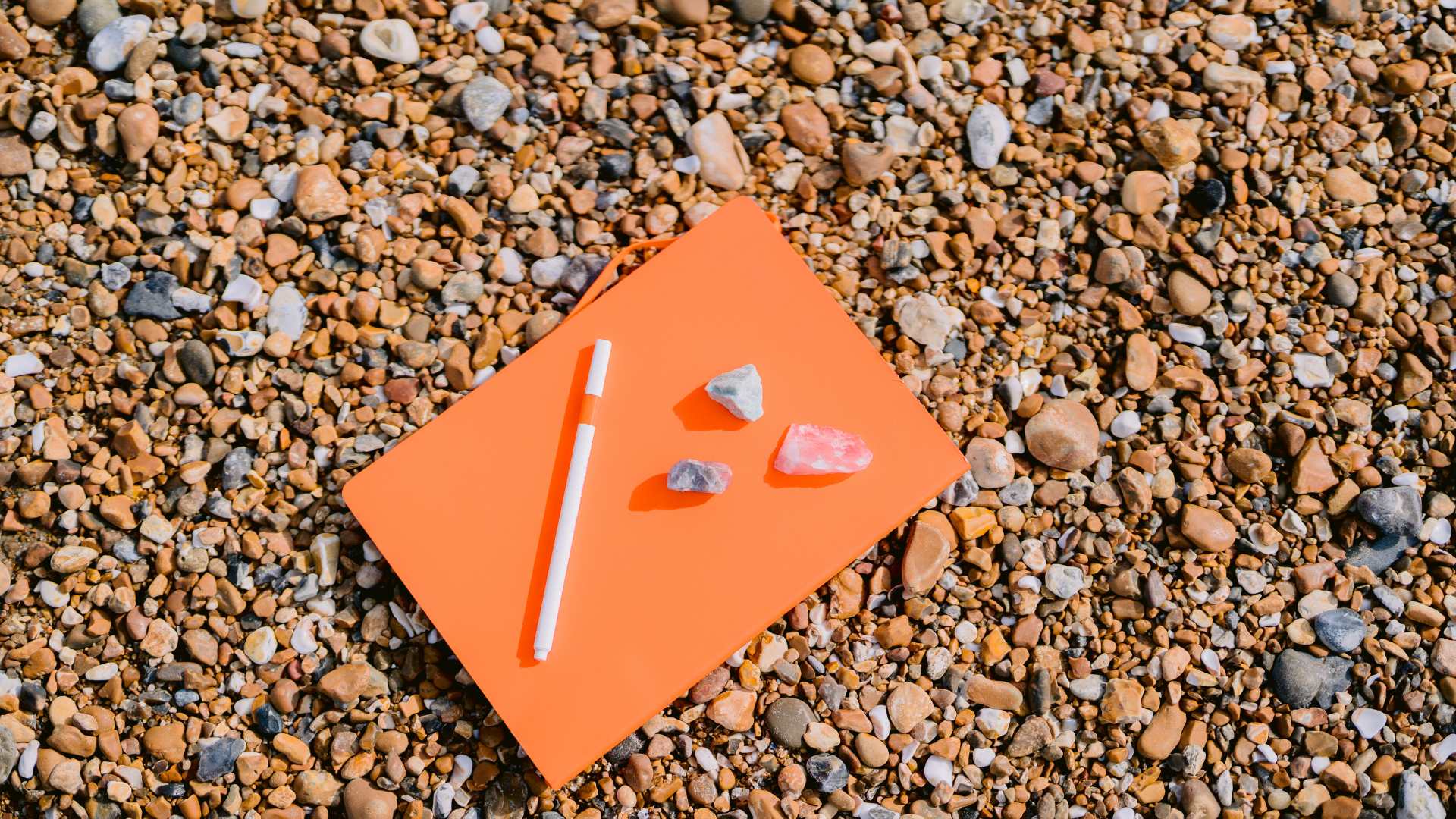 An orange notebook on a pebble beach with a pen and crystals. The notebook has been placed ready for someone to write and create a funeral.