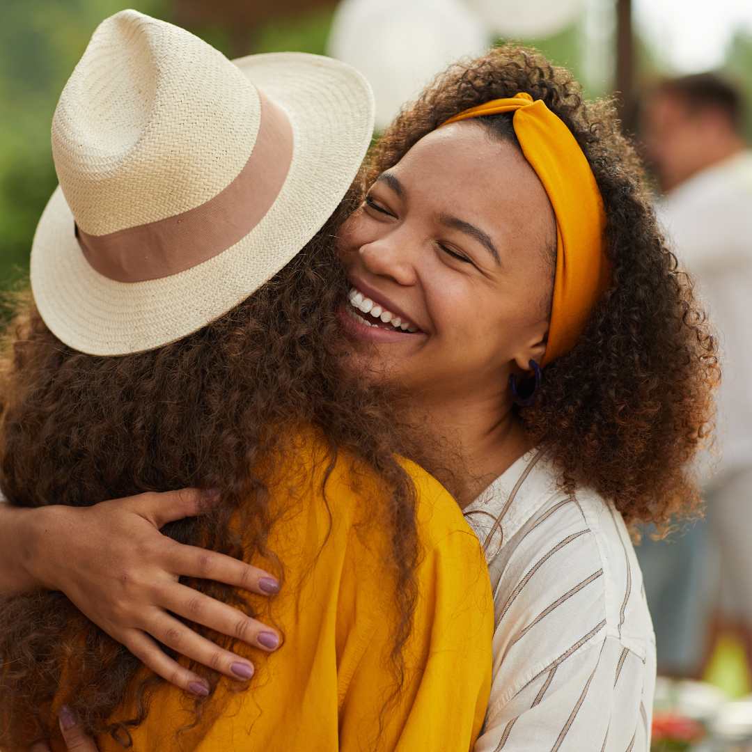 Two people hugging. both are wearing yellow. One has a face to the camera and is a person of colour with a yellow headband.